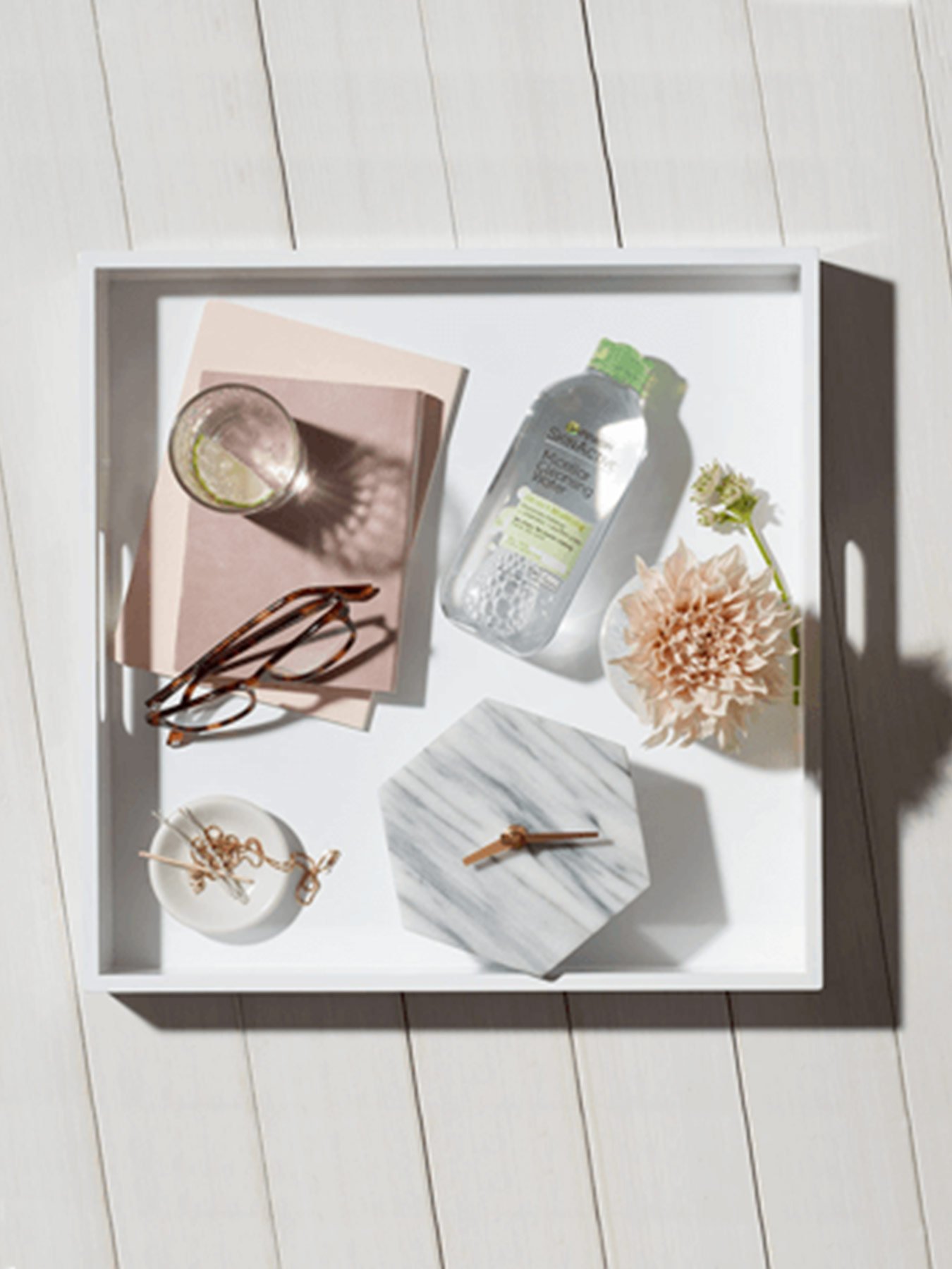 Garnier SkinActive Micellar Cleansing Water All-in-1 Mattifying in a white box with pink stationary, a lime slice in a clear glass, reading glasses, a white bowl of gold jewelry, a white marble octagonal clock with bronze hands reading 8:17, a white vase with a pink flower, and a small flower lying flat all on a white wooden background.